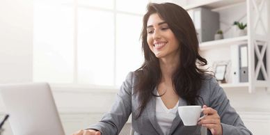 A woman smiling while working on a laptop and holding a coffee cup.