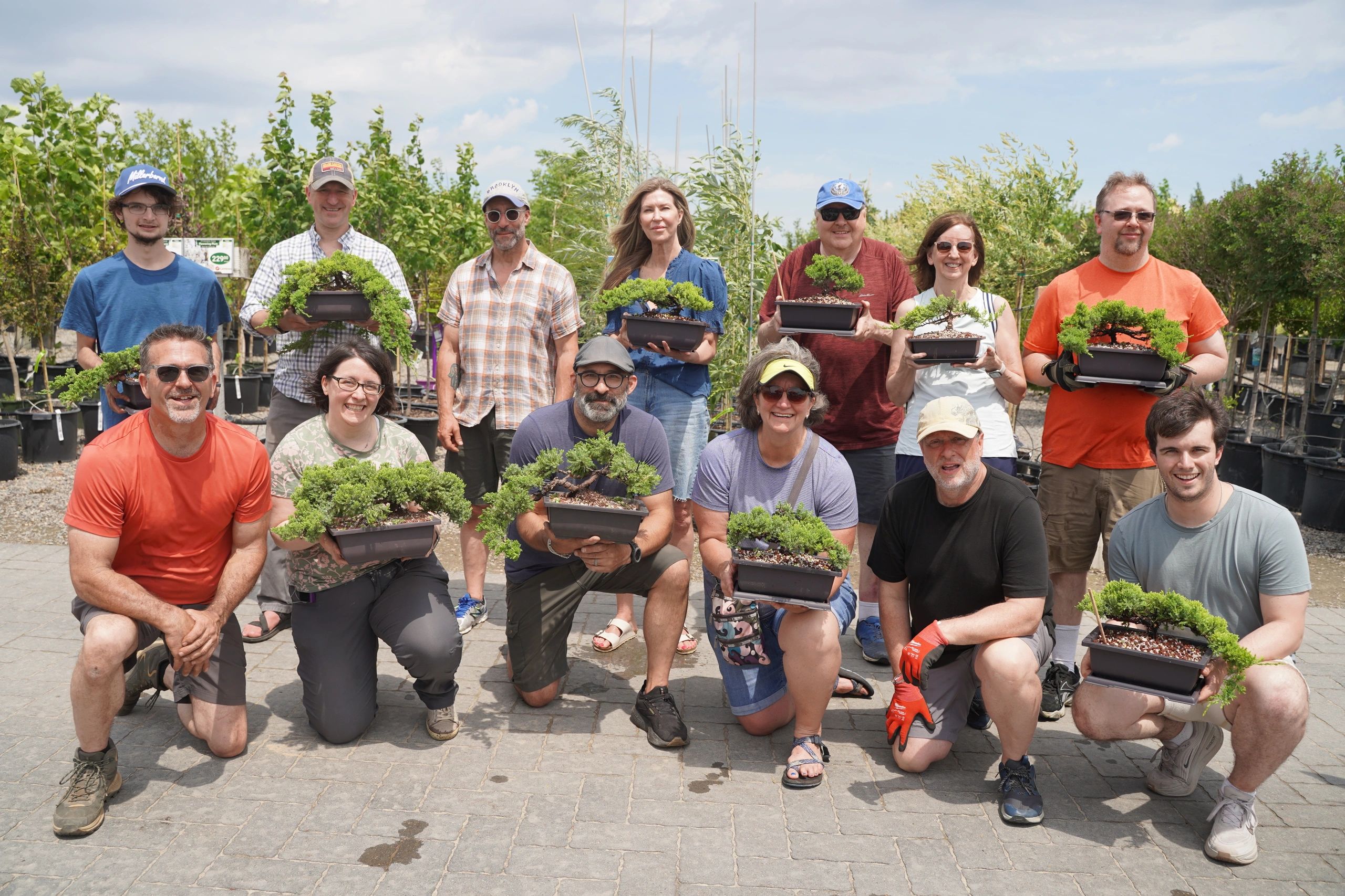 Group of people proudly holding bonsai trees outdoors on a sunny day.