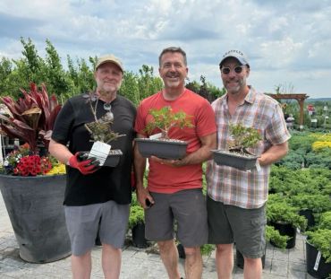 Three men happily holding bonsai plants at a garden center.