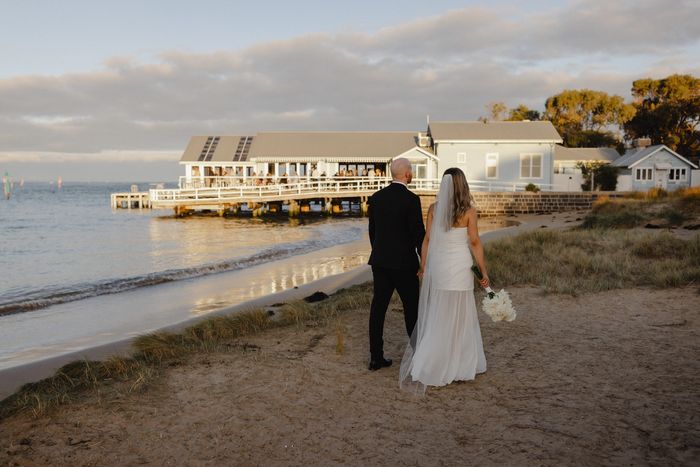 A newlywed couple walks along a beach toward a pier at sunset.