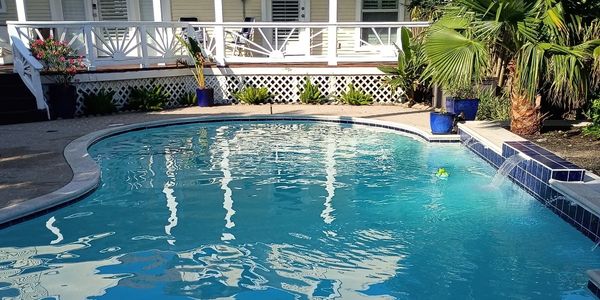 A clear pool in a backyard with a white deck and tropical plants.