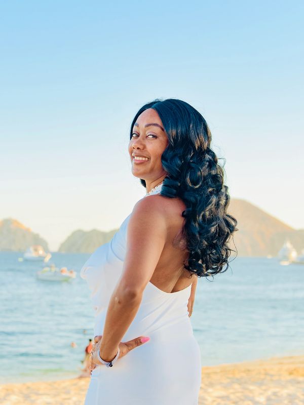 Woman in a white dress smiling on a beach with boats and hills in the background.