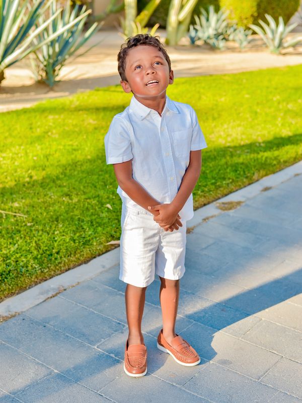 Young boy in white outfit standing on a sunny path looking up.