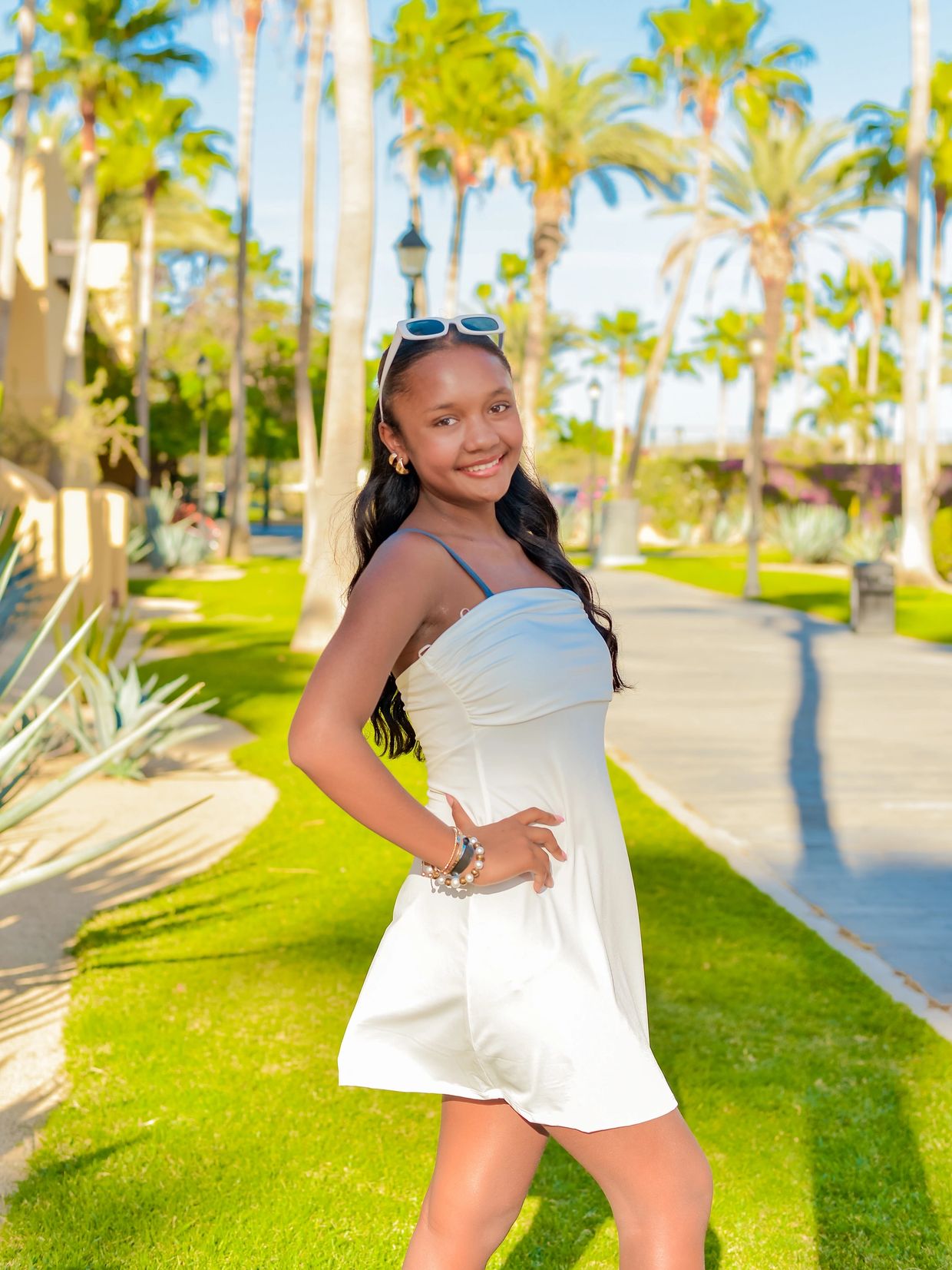 A young woman in a white dress smiles outdoors with palm trees in the background.