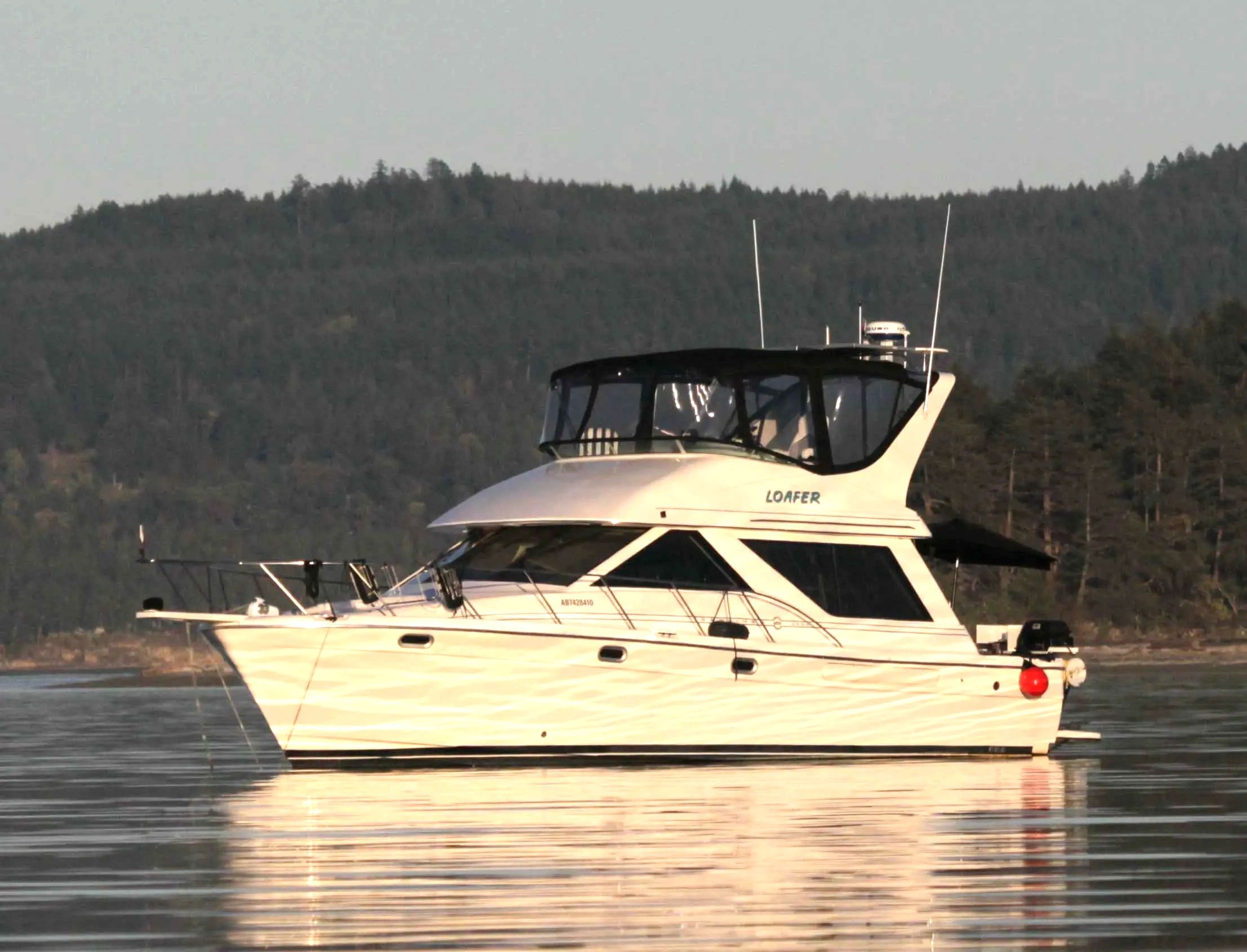 White yacht named 'LOAFER' floating on calm water near forested shore.