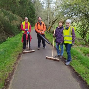 Four volunteers sweep a paved path in a green outdoor area.