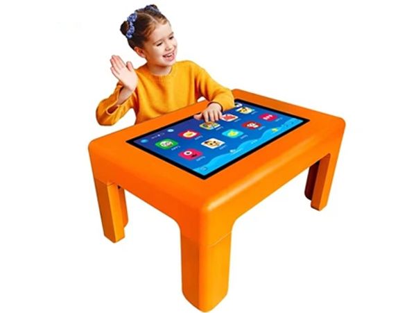 Young girl using an interactive orange touchscreen table, smiling and waving.