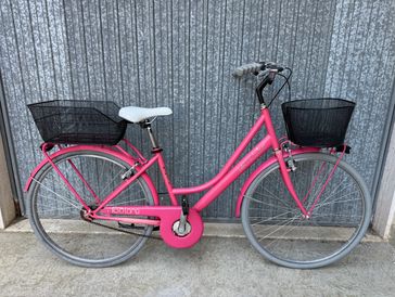 Pink bicycle with front and rear black baskets against a corrugated metal wall.