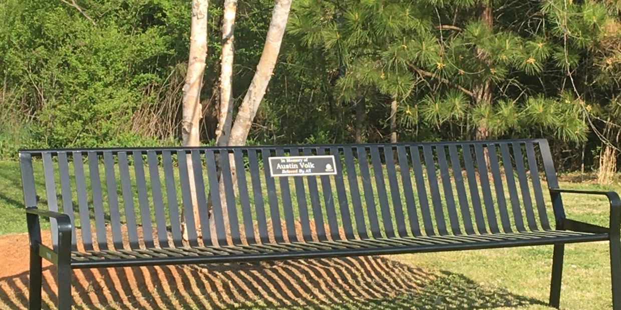 A black metal park bench with a dedication plaque in a green outdoor setting.