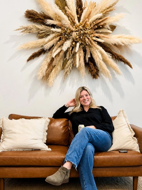 Woman sitting on a brown leather couch under a large decorative dried flower arrangement.