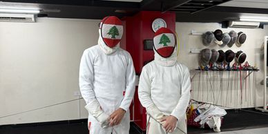 Two fencers wearing Lebanese flag face masks and white fencing gear.