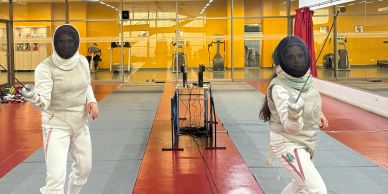 Two people fencing indoors in a gym with yellow walls and mirrors.
