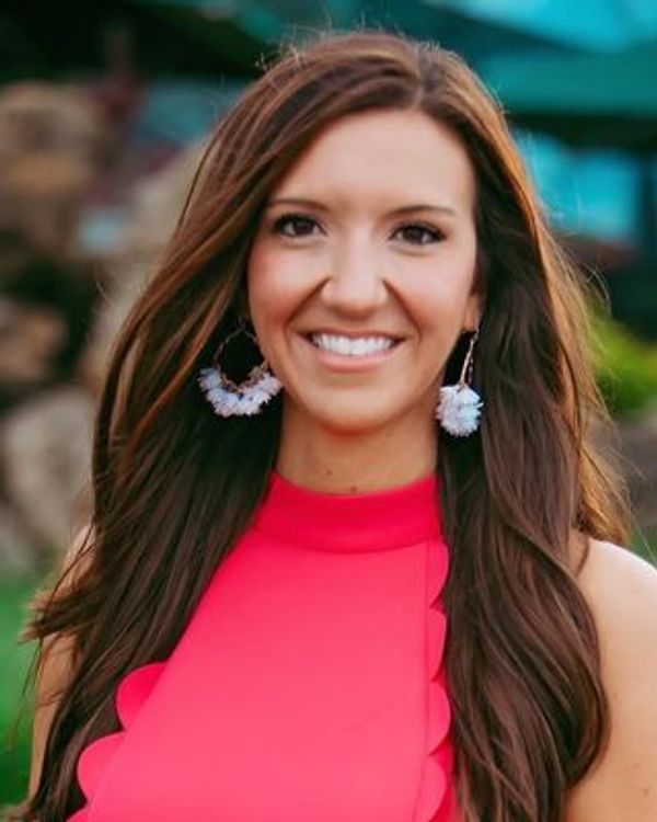 Smiling woman with long brown hair and white earrings in a bright pink top.