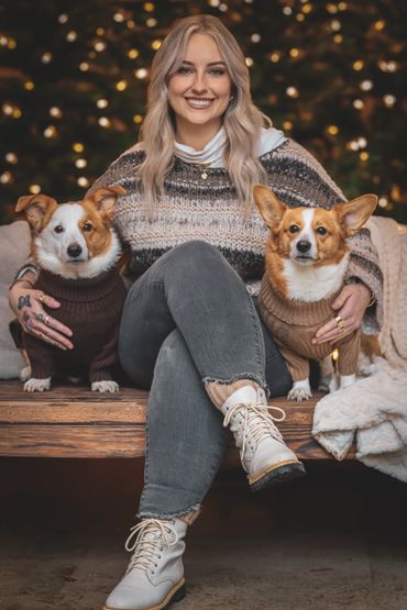 Woman sitting on a bench with two corgis in sweaters.