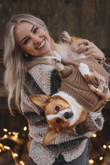 A smiling woman holds a corgi dog wearing a cozy sweater.