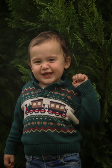 Smiling toddler in a green sweater with train design outdoors.