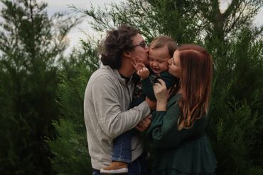 Parents kissing their smiling baby outdoors among green trees.