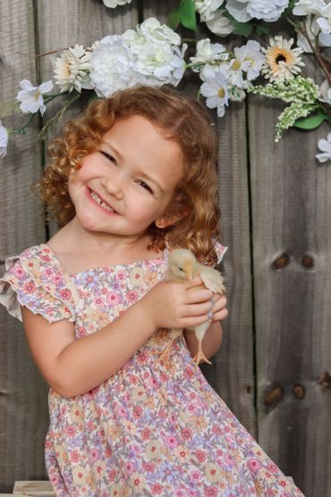 Smiling girl holding a chick with flowers behind her.