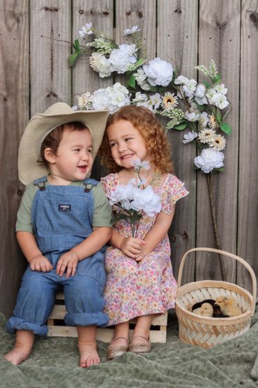 Two young children smiling, sitting with flowers and a basket of chicks.