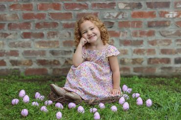 A little girl in a floral dress sitting on grass surrounded by purple tulips.