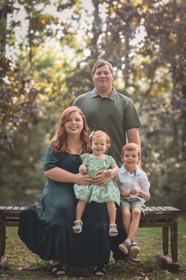 A family of four posing outdoors on a bench with a blurred forest background.