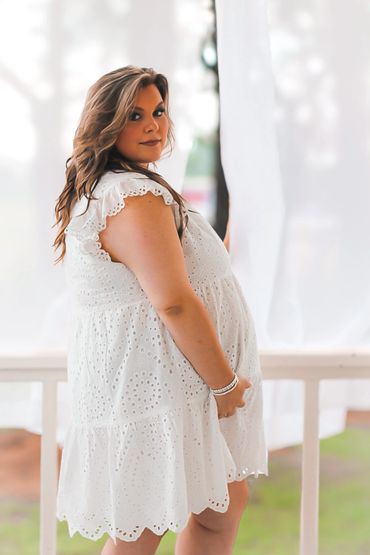 Pregnant woman in a white dress standing outdoors, gently holding her belly.