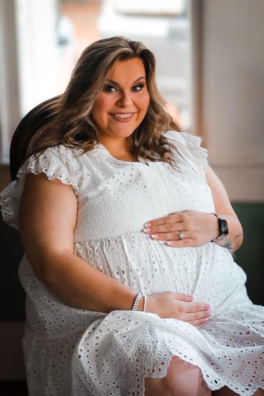 Pregnant woman in a white dress smiling and sitting indoors.