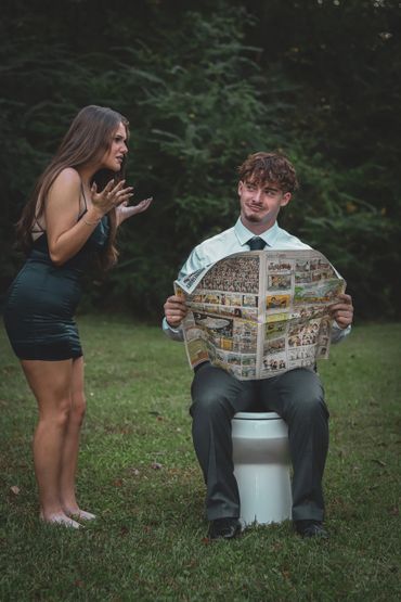 A man sits on a toilet outdoors reading a newspaper while a woman argues beside him.