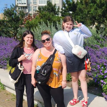Three women posing happily in front of purple flowers in an urban park on a sunny day.