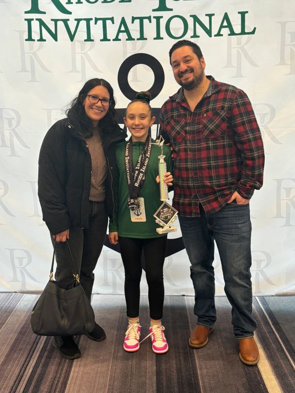 A young girl holding a trophy with two adults, all smiling at a Rhode Island Invitational event.