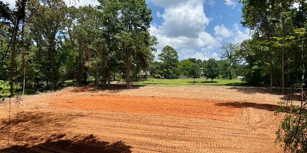 Cleared and leveled dirt lot surrounded by trees under a partly cloudy sky.