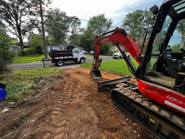 Excavator working on cleared ground near a truck and houses.