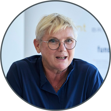 Woman with short blonde hair and glasses speaking, wearing a navy blue top.
