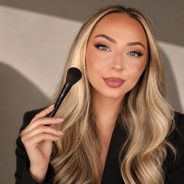 Woman with blonde hair holding a makeup brush, smiling confidently.