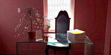 Elegant glass desk with vintage chair by a tall window in a softly lit room.