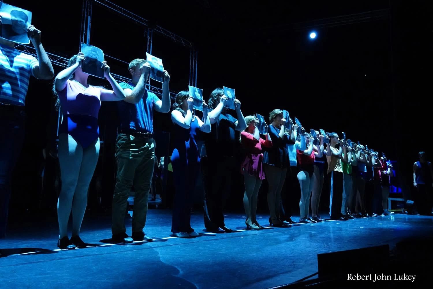 Performers on stage holding photos in front of their faces under blue lighting.