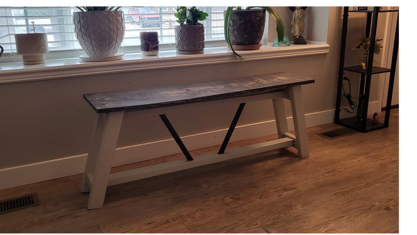Rustic wooden bench with a dark top and white legs beneath a window with plants.