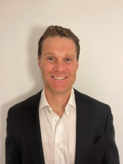 Smiling man in a black suit jacket and white shirt against a plain background.
