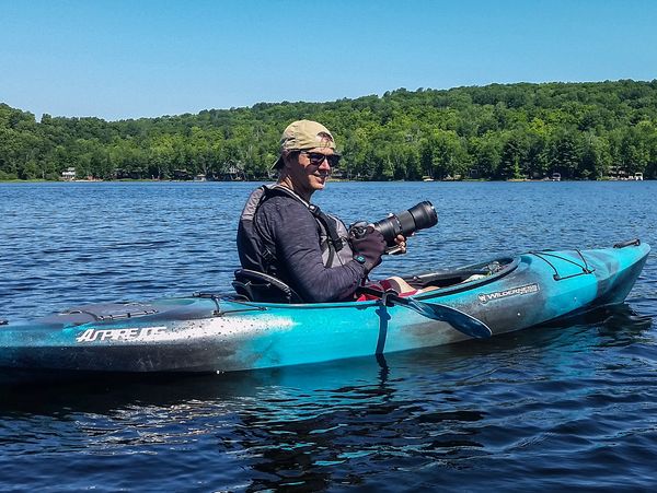 Man in a blue kayak holding a camera on a sunny lake day.