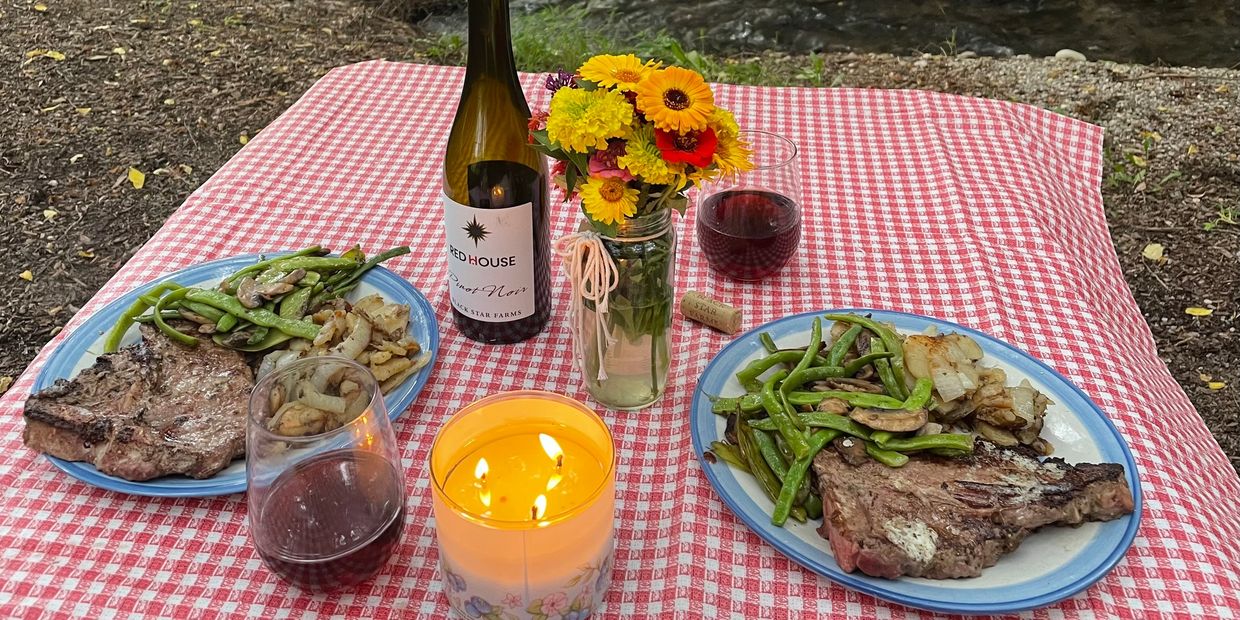 Outdoor picnic with steak, green beans, potatoes, red wine, and a candle on a checkered cloth.
