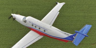 A white and blue Royal Flying Doctor Service plane flying over green fields.