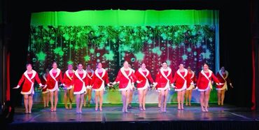 Dancers in red and white Santa outfits perform on stage with festive green backdrop.