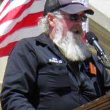 Bearded man in sunglasses speaking at a podium with an American flag behind him.