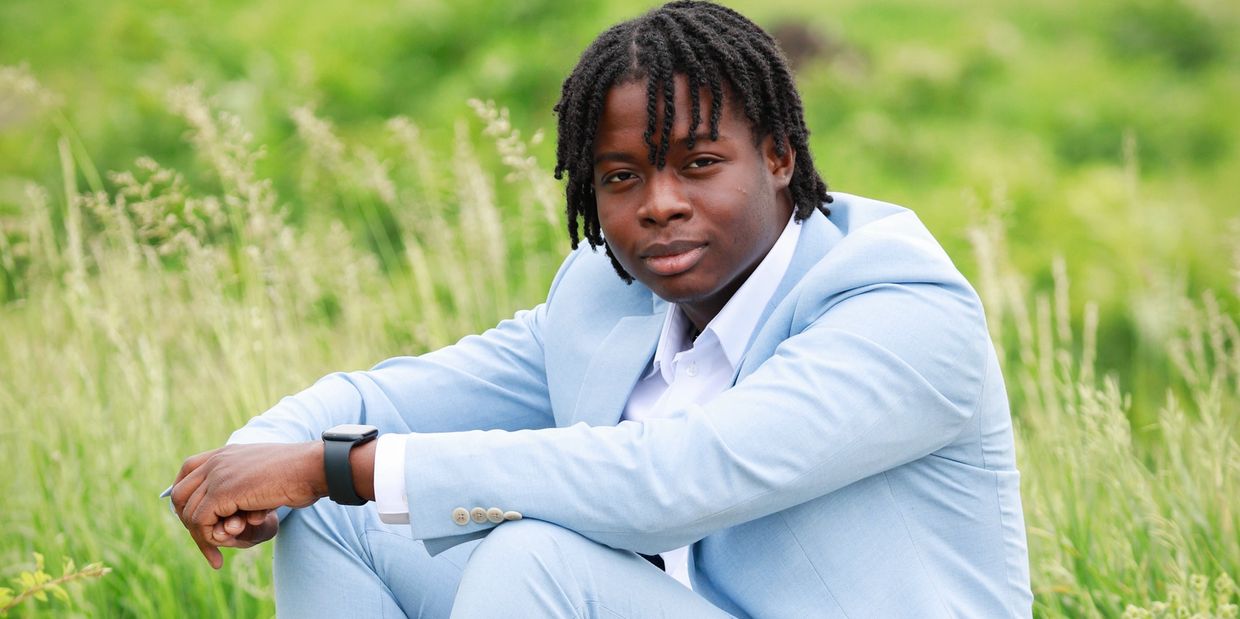 Young man in a light blue suit sitting in a grassy field.