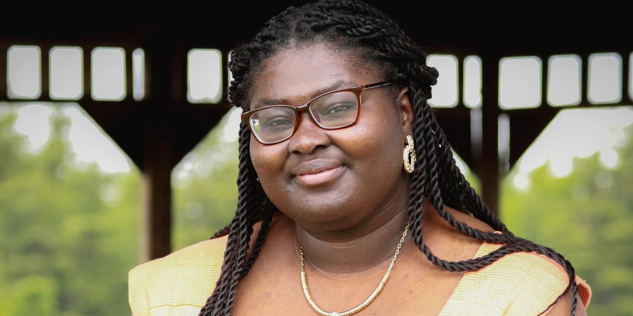A smiling woman with braided hair and glasses, wearing a yellow top and gold jewelry.