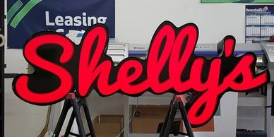 Bright red 'Shelly's' sign in a workspace with tools and leasing sign.