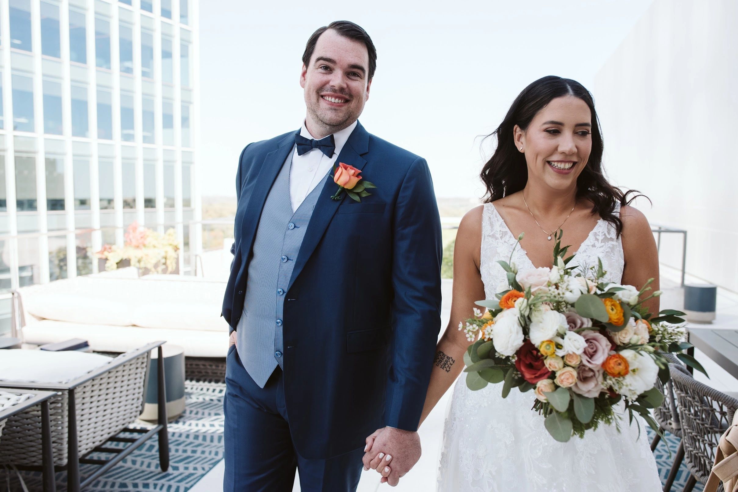 Smiling bride and groom holding hands on a sunny balcony.