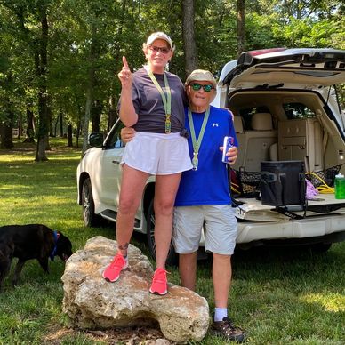 Two people celebrating outdoors with medals and a dog nearby.