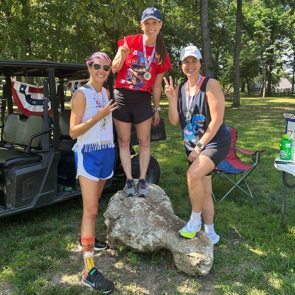 Three women posing with medals outdoors on a sunny day.