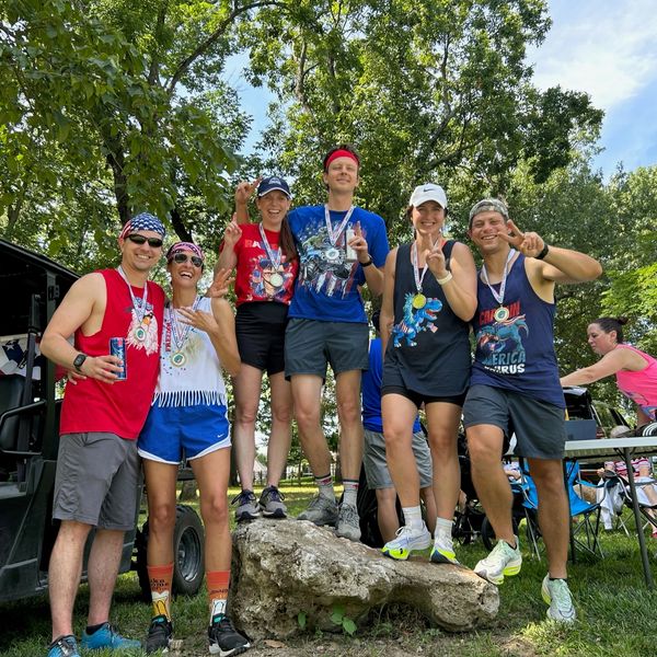 Group of six smiling runners posing with medals outdoors.